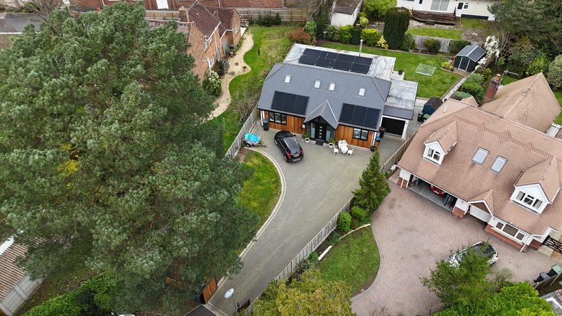 Aerial view of a modern chalet-style house with black solar panels on two roof faces and a flat-roof array